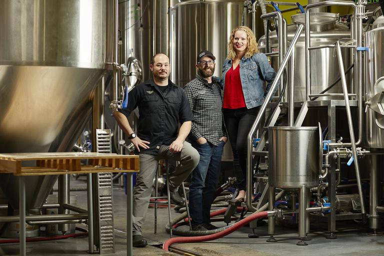 a woman and two men surrounded by brewery equipment
